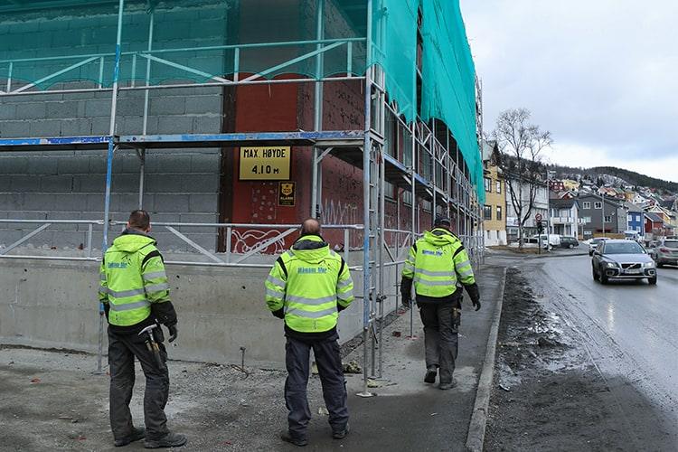 Tre mann i arbeidsklær foran et bygg med stillas